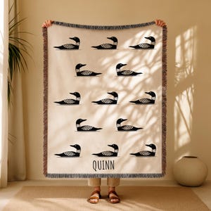 Peut inclure: Une couverture tissée beige avec un motif répété d'illustrations de huards noirs et blancs. La couverture a un bord à franges et le nom "QUINN" est imprimé en noir en bas. La couverture est tenue par une personne.