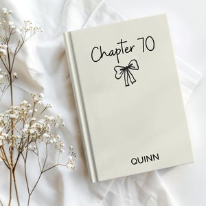 May include: A cream-colored book with the text "Chapter 70" and a bow illustration in black. The name "QUINN" is printed at the bottom. The book is on a white fabric surface, with sprigs of white flowers.