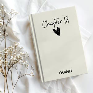 Pode incluir: Um livro creme com o texto "Chapter 18" e uma ilustração de coração preto na capa. O nome "QUINN" está impresso na parte inferior. O livro está sobre um fundo branco texturizado com delicadas flores brancas.