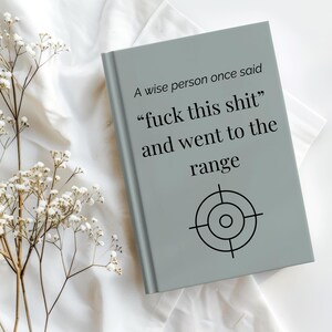 Puede incluir: Un cuaderno gris con el texto "A wise person once said 'fuck this shit' and went to the range" y un símbolo de diana. El cuaderno está sobre una superficie blanca con flores secas.