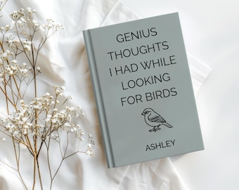 May include: A light gray book with the text "GENIUS THOUGHTS I HAD WHILE LOOKING FOR BIRDS" and a bird illustration. The name "ASHLEY" is printed at the bottom. White flowers are in the background.