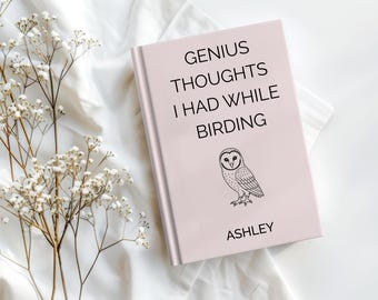 May include: A light pink book with the title "GENIUS THOUGHTS I HAD WHILE BIRDING" and an illustration of an owl. The author's name, Ashley, is printed at the bottom. The book is on a white surface with sprigs of white flowers.