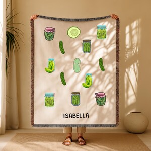 May include: A beige throw blanket with a pattern of cartoon pickles and jars. The design includes whole pickles, pickle slices, and jars with pink, blue, and brown lids. The name "ISABELLA" is printed in black block letters at the bottom.