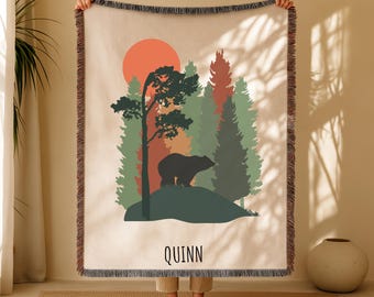 May include: A beige throw blanket with a forest scene featuring a bear, trees, and a setting sun. The design includes the name "Quinn" at the bottom. The blanket has a fringed edge.