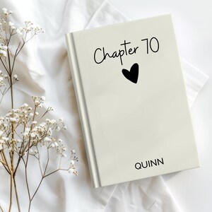 May include: A cream-coloured book with the text "Chapter 70" and a black heart symbol. The name "Quinn" is printed at the bottom. The book is on a white cloth with sprigs of white flowers.