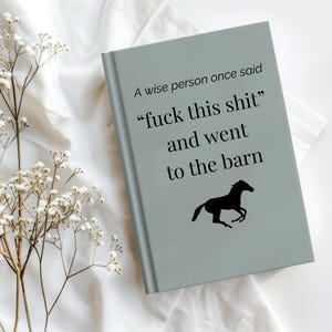 May include: A gray journal with the text "A wise person once said 'fuck this shit' and went to the barn" and a black horse silhouette. The journal is on a white surface with white flowers.