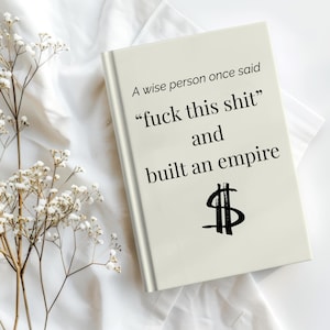 Puede incluir: Un libro de tapa dura con el texto "A wise person once said 'fuck this shit' and built an empire" en fuente negra. Un símbolo de dólar está debajo. El libro está sobre una superficie blanca con flores blancas.