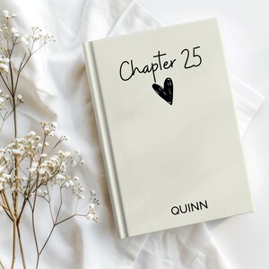 May include: A cream-colored book with the text "Chapter 25" and a black heart illustration. The name "Quinn" is printed at the bottom. The book is on a white cloth with sprigs of white flowers.