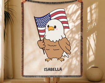 May include: A beige throw blanket featuring a cartoon bald eagle holding an American flag. The eagle has brown and white feathers, a yellow beak, and pink cheeks. The name "ISABELLA" is printed in black below the eagle.