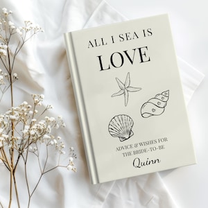 May include: A cream-colored book with the text "ALL I SEA IS LOVE" and seashell illustrations. The book includes the text "ADVICE & WISHES FOR THE BRIDE-TO-BE" and the name "Quinn". The book is on a white cloth.