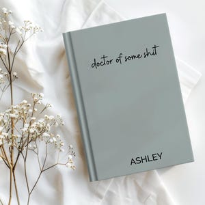 May include: A grey notebook with the text "doctor of some shit" and the name "ASHLEY" in black script. The notebook is on a white surface with delicate white flowers.