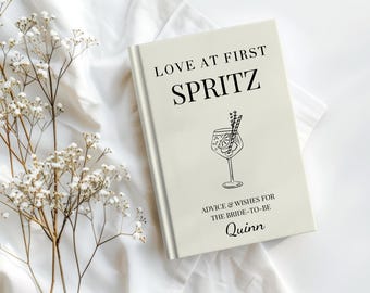 May include: A cream-colored book with the text "LOVE AT FIRST SPRITZ" and a line drawing of a cocktail glass. Below the title is "ADVICE & WISHES FOR THE BRIDE-TO-BE" and the name "Quinn". The book is on a white surface with white flowers.