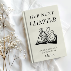 May include: A cream-colored book with the text "HER NEXT CHAPTER" in black. Below the title is a floral illustration and the text "ADVICE & WISHES FOR THE BRIDE-TO-BE Quinn". The book is on a white surface with white flowers.