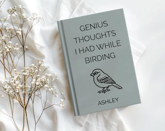 May include: A light gray book with the text "GENIUS THOUGHTS I HAD WHILE BIRDING" and a bird illustration. The name "ASHLEY" is printed at the bottom. The book is on a white surface with white flowers.