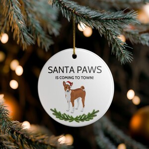 May include: A white, round Christmas ornament with the text "SANTA PAWS IS COMING TO TOWN!" features a Basenji dog wearing a Santa hat. The ornament is decorated with a green garland and a gold string for hanging. The background is a blurred Christmas tree.