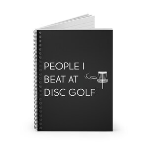 May include: A black spiral-bound notebook with the text "PEOPLE I BEAT AT DISC GOLF" in white. A white graphic of a disc golf basket and flying disc is also on the cover. The notebook is open.