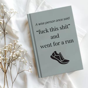 Może przedstawiać: Szary notatnik z napisem "A wise person once said 'fuck this shit' and went for a run" i czarną grafiką buta do biegania. Notatnik leży na białej powierzchni z białymi kwiatami.
