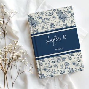 May include: A hardback book with a cream-coloured cover featuring a dark blue floral pattern. A solid dark blue horizontal band across the centre displays the white text "chapter 70" in a script font and the name "ASHLEY" in a sans-serif font.