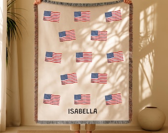 May include: A beige throw blanket featuring multiple American flags in red, white, and blue. The blanket has a fringed edge and the name "ISABELLA" printed in black. The blanket is being held up by a person.