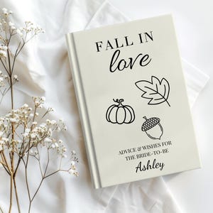 May include: A cream-colored hardcover book with the text FALL IN LOVE, ADVICE & WISHES FOR THE BRIDE-TO-BE, and the name Ashley printed in black. The cover features black line art illustrations of a pumpkin, a leaf, and an acorn.