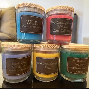 May include: Five glass candle jars with wooden lids. The candles are in various colors, including blue, red, purple, yellow, and green. Each jar has a brown leather label with text. The text on the labels reads: "WTF... What's the Function", "The Calming Scent of Proper Data Collection", "Smells Like Worlds Best Behavior Therapist", "Behavior Analyst Vibes", and "Smells Like Positive Reinforcement In Here".
