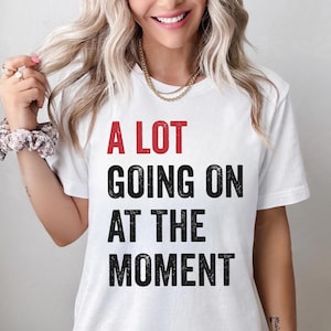 以下が含まれることがあります： 白地に「A LOT GOING ON AT THE MOMENT」という文字が黒と赤で書かれたTシャツです。