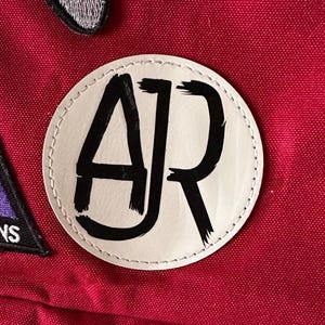 May include: A red backpack with a white circle patch that says "AJR" in black lettering. The backpack also has a gray video game controller patch and a colorful triangle patch with the text "Imagine Dragons".