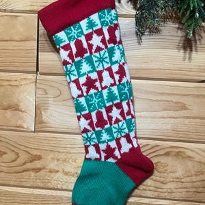 May include: A Christmas stocking with a red cuff and toe, and a green heel. The body of the stocking is white with a pattern of red stars, green Christmas trees, and green snowflakes. The stocking is hanging on a wooden surface.