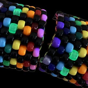 Puede incluir: Dos pulseras kandi coloridas, cada una tejida con un patrón de cuentas negras, moradas, azules, verdes, amarillas, naranjas, rojas y rosas. Algunas cuentas brillan. Las pulseras están conectadas por un hilo verde.