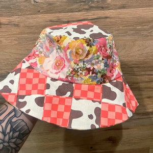 May include: Reversible bucket hat with a patchwork design. One side features a floral pattern in pink, yellow, and purple. The other side has a cow print and a red and white checkered pattern. The hat is made of fabric.