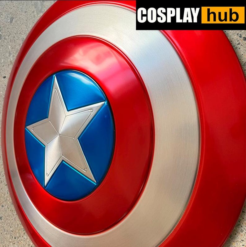 Pu&ograve; includere: Uno scudo di Captain America con una combinazione di colori rosso, bianco e blu. Lo scudo presenta una stella argentata in un cerchio blu al centro, circondata da bande rosse e argentate concentriche. Il testo "COSPLAY hub" &egrave; nell'angolo in alto a destra.