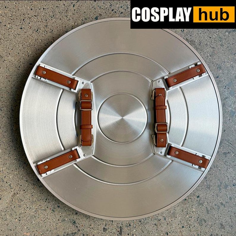 Pu&ograve; includere: Uno scudo circolare argentato con cinghie e fibbie in pelle marrone. Lo scudo ha una finitura in metallo spazzolato e un design circolare centrale. Il testo "COSPLAY hub" &egrave; visualizzato in alto a destra.