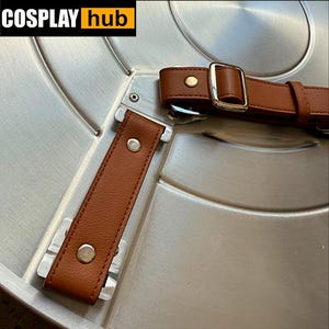 Pu&ograve; includere: Primo piano di uno scudo color argento con cinturini in pelle marrone e fibbie argentate. Lo scudo ha un design circolare con creste concentriche. Il testo "COSPLAY hub" &egrave; nell'angolo in alto a sinistra.