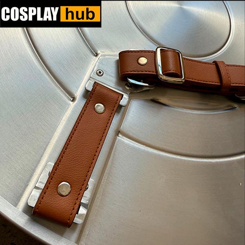 Pu&ograve; includere: Primo piano di uno scudo color argento con cinturini in pelle marrone e fibbie argentate. Lo scudo ha un design circolare con creste concentriche. Il testo "COSPLAY hub" &egrave; nell'angolo in alto a sinistra.