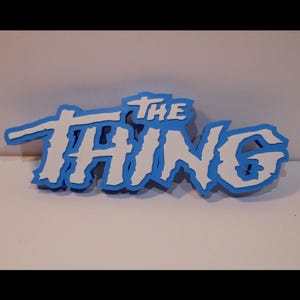 Może przedstawiać: Niebiesko-biały znak z napisem "THE THING" w stylizowanej czcionce. Tekst jest biały z niebieskim obrysem, na białym tle. Znak jest prawdopodobnie przedmiotem dekoracyjnym lub rekwizytem.