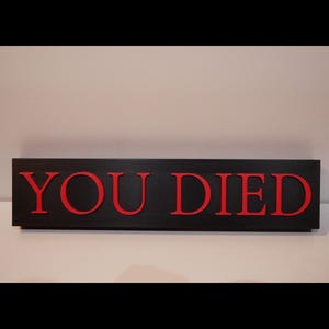 May include: A rectangular black sign with the words "YOU DIED" in red, bold letters. The sign is likely made of wood or a similar material, and the letters are raised, creating a 3D effect. The background is a plain white surface.