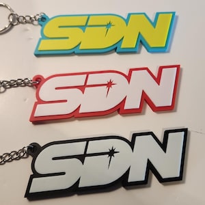 May include: Three keyrings with the letters "SDN" in a bold, stylised font. The top keyring has yellow letters with a blue outline, the middle one has red letters, and the bottom one has white letters with a black outline. Each keyring has a silver keyring.