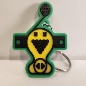 May include: A green, yellow, and black keychain with a unique design. The keychain features a yellow face with a black triangle and two black squares. A green handle with a black circle is attached to the top. A silver key ring is attached.