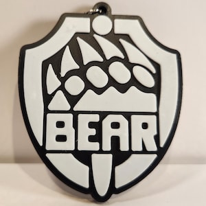 May include: A black and white shield-shaped keychain featuring a graphic of a bear paw print at the top and the word BEAR in bold, capital letters at the bottom. The design is rendered in a high-contrast, two-tone color scheme with a small metal ring.