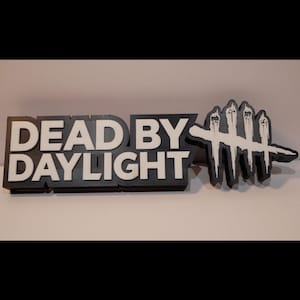 Op de afbeelding: Een zwart-wit bord met de tekst "DEAD BY DAYLIGHT" in vette blokletters. Het bord bevat ook een wit handsymbool met vier vingers. Dit is waarschijnlijk een decoratieartikel of merchandise.