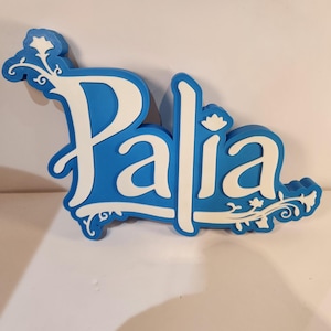 May include: A blue and white decorative sign with the name "Palia" in a stylized font. The sign has a blue background with white lettering and decorative floral accents. The sign is made of a thick material.