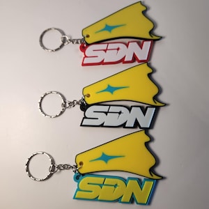 May include: Three keychains with the letters "SDN" in white, red, black, and blue. Each keychain has a yellow fin-shaped design with a blue starburst. The keychains are attached to silver keyrings.