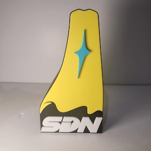 Puede incluir: Objeto decorativo amarillo y negro con un diseño de estrella azul y las letras "SDN" en blanco. El objeto tiene una forma única y se mantiene erguido.