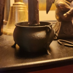 Könnte beinhalten: Ein schwarzer Kessel aus Gusseisen mit drei Beinen und einem Griff. Der Kessel steht auf einer dunklen Holzoberfläche. Eine braune Kerze steht auf dem Kessel.
