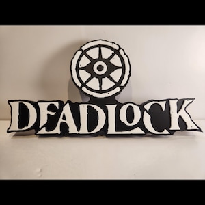 Op de afbeelding: Een zwart-wit bord met het woord "DEADLOCK" in vette, gestileerde letters. Boven het woord staat een cirkelvormig embleem met een wielachtig ontwerp. Het bord is waarschijnlijk een decoratief item of logo.