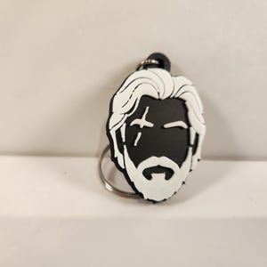 May include: A black and white keyring featuring a stylised graphic of a person with wavy hair, a full beard, and a distinct scar over one eye. The design is rendered in a flat, high-contrast style with a metal ring attached at the top.