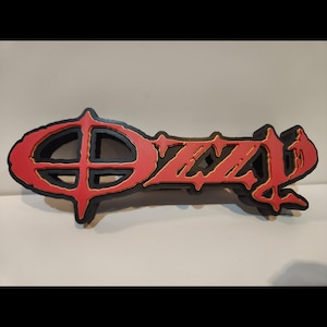 May include: A red and black logo with the name "Ozzy" in a distinctive font. The logo has a black outline with a gold trim, and the letters are in a bold, red colour. This is likely a piece of merchandise associated with the musician Ozzy Osbourne.