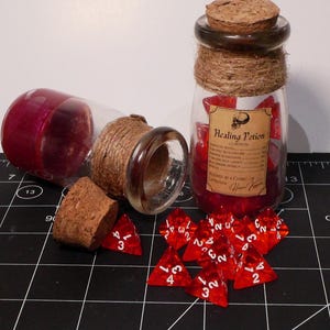 May include: Two clear glass bottles with cork stoppers and twine-wrapped necks. One bottle stands upright, filled with red translucent polyhedral dice and a label reading Healing Potion. The other bottle lies on its side with a loose cork and red dice.