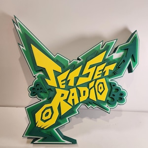 May include: A layered, three-dimensional sign featuring the text "JET SET RADIO" in yellow, outlined in green. The sign has a graffiti-style design with sharp angles and a white border. The background is a plain white.