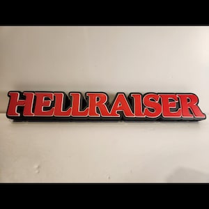 May include: A rectangular sign featuring the word "HELLRAISER" in red, bold lettering with a white border, set against a black background. The sign is likely made of a solid material.